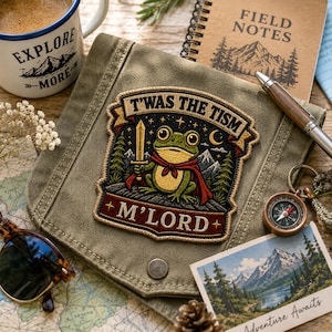 May include: An embroidered patch featuring a green frog wearing a red cape and holding a sword, set against a dark starry night with mountains and trees. The text reads T'WAS THE TISM and M'LORD, displayed on a folded olive green canvas fabric.