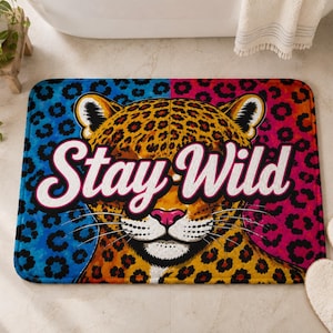 May include: A rectangular bath mat featuring a central illustration of a leopard face with a yellow, black, and brown spotted coat. The background is split into blue and pink leopard print sections. The white text Stay Wild is centered.