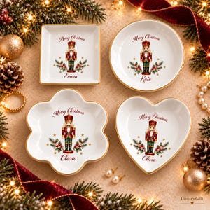 May include: Four white ceramic trinket dishes with gold rims, featuring a nutcracker illustration, holly sprigs, and the text Merry Christmas. Each dish is personalized with a name: Emma, Kate, Clara, or Olivia, in different shapes.