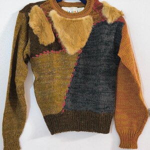 Puede incluir: Un suéter de punto de manga larga con un diseño de parches en tonos amarillo mostaza, marrón oscuro y gris carbón. La prenda incluye parches de piel sintética texturizada en el pecho y los hombros, acentuados por costuras visibles en color rosa.
