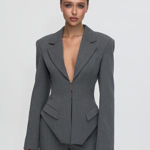 May include: A charcoal gray blazer featuring a deep V-neckline, structured shoulders, and long sleeves. The garment has a single hook-and-eye closure at the center front, two decorative flap pockets at the waist, and a pointed, asymmetrical hemline.