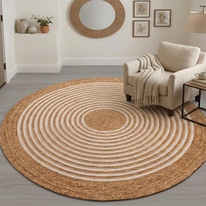 Può includere: Un tappeto rotondo con un motivo a cerchi concentrici in fibre di juta naturale color marrone chiaro e bianco. Il tappeto presenta una struttura intrecciata e testurizzata con bande alternate di marrone chiaro e bianco, creando un design circolare.