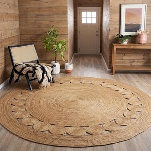 Può includere: Un grande tappeto rotondo in juta color naturale caratterizzato da una trama a spirale centrale circondata da un bordo decorativo composto da dischi circolari in juta. Il tappeto è posizionato su un pavimento in legno chiaro in una stanza.