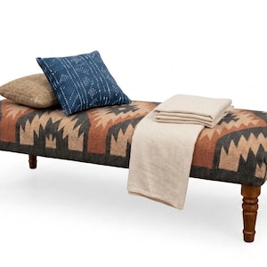 May include: A rectangular upholstered bench with four turned wooden legs, featuring a geometric pattern in shades of brown, black, and tan. The bench is topped with a blue patterned square pillow, a tan pillow, and a folded cream-colored throw blanket.
