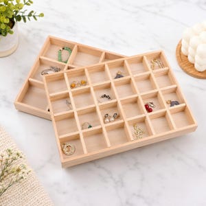 May include: Two light-colored wooden jewelry organizer trays with multiple square compartments. The trays are arranged in an overlapping pattern and contain various small pieces of jewelry, including rings, earrings, and a green beaded bracelet.