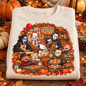 Op de afbeelding: Een crèmekleurig sweatshirt met een grafische illustratie van horrorfilmfiguren en Snoopy die bakken in een pompoenbakkerij. Het ontwerp bevat herfstbladeren, pompoenen, taarten en de tekst PUMPKIN BAKERY in een feestelijk herfstthema.