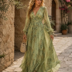 Puede incluir: Un vestido largo de color verde salvia con escote en V, mangas largas transparentes tipo obispo con puños y cintura fruncida. La tela tiene un estampado floral en tonos amarillos y marrones. Se sostiene un pequeño bolso de paja en la mano.