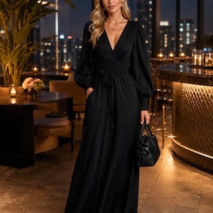 May include: A floor-length black dress with a deep V-neckline, long bishop sleeves with fitted cuffs, and a smocked elastic waist featuring a tie-front detail. The dress has a pleated texture throughout and includes side pockets, paired with a black handbag.