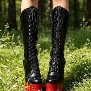 May include: A pair of knee-high black boots featuring a lace-up front with silver eyelets, a side zipper closure, and a thick, solid red platform sole. The boots have a smooth, glossy finish and extend to the lower calf area.