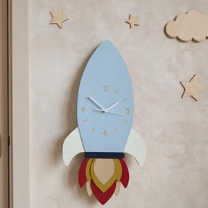 May include: A rocket-shaped wall clock in light blue with white hands and numerals 3, 6, 9, and 12. The base features white fins and a flame design in red, gold, and natural wood. Surrounding the clock are wooden cloud and star wall decorations.