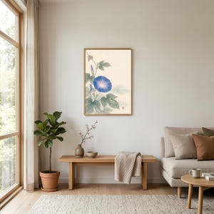 May include: A framed wall art print featuring a botanical illustration of a single vibrant blue morning glory flower with green leaves and vines on a light cream background, displayed in a simple wooden frame on a plain white wall.