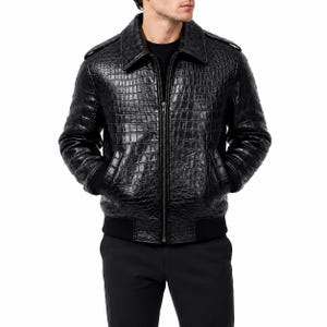 Puede incluir: Una chaqueta negra con un patrón texturizado de piel de cocodrilo, cremallera frontal completa, cuello en punta y puños y dobladillo acanalados en negro. La prenda se lleva sobre una camisa negra lisa y se combina con pantalones negros.