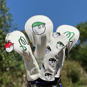 Könnte beinhalten: Ein Set weißer Golfschlägerhauben mit gestickten grünen Mützen auf Golfbällen, einem grünen kursiven M-Logo und einem kleinen Golfball-Anhänger mit roter Mütze. Die Hauben zeigen den Schriftzug Malbon Golf und ein wiederkehrendes Golfballmuster.