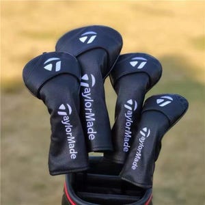 Könnte beinhalten: Vier schwarze Golfschlägerhauben aus strukturiertem Material, jeweils mit dem weißen TaylorMade-Logo und dem vertikal aufgedruckten Markennamen. Die Hauben sind zusammen angeordnet und teilweise in eine schwarze Golftasche mit rotem Rand gesteckt.
