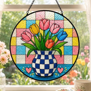 May include: A circular stained glass suncatcher featuring a bouquet of pink, yellow, orange, and blue tulips in a blue and white checkered vase. The background consists of a colorful grid pattern with two small pink hearts at the base.