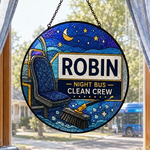 May include: A circular stained glass suncatcher featuring a night sky with a crescent moon and stars. The design depicts a bus seat and a broom with the text ROBIN NIGHT BUS CLEAN CREW. The piece is suspended by a black metal chain.