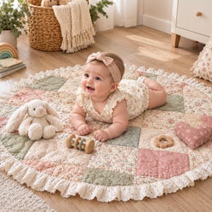 May include: A round, quilted baby play mat featuring a patchwork design in muted sage green, dusty pink, and cream floral patterns, finished with a white lace-trimmed ruffled edge. A plush cream-coloured bunny toy and wooden rattle are included.