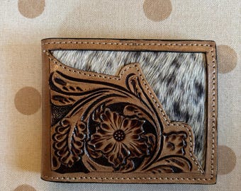 May include: A bi-fold wallet featuring a combination of tan leather with intricate floral tooling and a section of black and white cowhide hair-on leather. The leather is stitched with light-colored thread along the edges of the wallet.