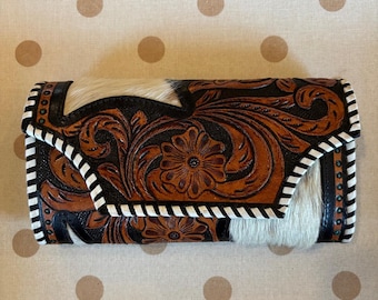 May include: A rectangular wallet featuring a combination of brown leather with intricate floral tooling and patches of white and brown cowhide. The edges are finished with a white and black whipstitch detail and small turquoise-colored studs.