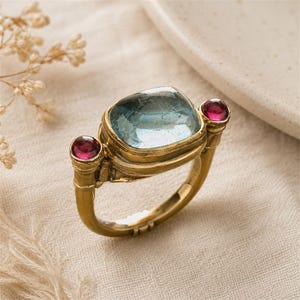 Könnte beinhalten: Ein goldener Ring mit einem großen, hellblauen Edelstein im Kissenschliff in der Mitte, flankiert von zwei kleineren, runden, dunkelrosa Edelsteinen auf beiden Seiten. Der Metallring hat eine texturierte, matte Oberfläche mit feinen Details.
