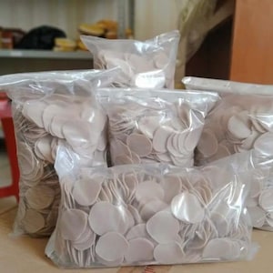 May include: Multiple clear plastic bags filled with small, round, off-white, flat, disc-shaped pieces. The bags are stacked and arranged to show a large quantity of these uniform, circular items, which appear to be thin, dry, and opaque in texture.