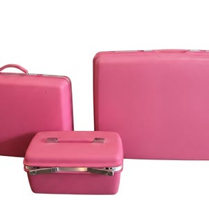 Puede incluir: Un conjunto de tres maletas rígidas de color rosa brillante y un maletín de aseo a juego. Las piezas de equipaje presentan superficies lisas de color sólido, formas rectangulares y herrajes de metal plateado, incluyendo asas y cierres.