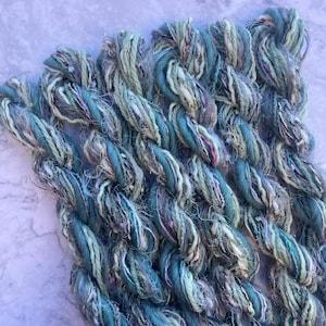 Puede incluir: Primer plano de varios hilos de lana hilada a mano en tonos turquesa, azul y verde. El hilo tiene un aspecto texturizado con hebras plateadas y rosas. El hilo está dispuesto en un patrón trenzado, adecuado para tejer u otras manualidades con fibra.