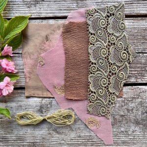 以下が含まれることがあります： ピンク、ブラウン、ベージュの色合いの生地見本のアソートメント。テクスチャード加工されたブラウンの生地と繊細なレースのトリムが含まれています。麻ひもの束と、緑の葉を持つピンクの花も、風化した木製の表面に配置されています。