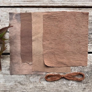 Może przedstawiać: Widok z góry przedstawia kolekcję brązowych, teksturowanych papierów i wiązkę brązowego sznurka. Papiery różnią się fakturą, w tym pogniecioną tkaniną, papierem w kropki i papierem z szorstkimi krawędziami. Paleta kolorów jest ziemista, z odcieniami brązu.