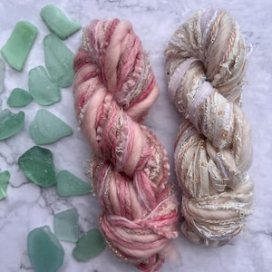May include: Two skeins of yarn in shades of pink, cream, and gold, with a textured, chunky appearance. The yarn is arranged next to several pieces of sea glass in varying shades of green, creating a visually appealing contrast.