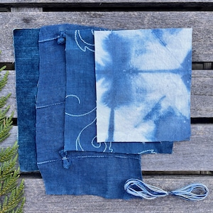 以下が含まれることがあります： さまざまな青色の濃淡で藍染めされた生地の見本。タイダイ柄の生地、白い刺繍が施された生地、青い糸の束が含まれています。風化した木製の表面に配置されています。