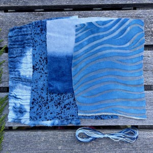 以下が含まれることがあります： さまざまな模様と質感の青い生地見本。タイダイ、斑点、波のようなデザインが含まれています。青と白の糸の束が下にあります。生地は風化した木製の表面に配置されています。