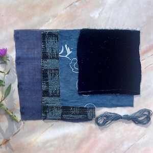 Pode incluir: Uma variedade de amostras de tecido tingidas com índigo em várias texturas e padrões. As amostras incluem um azul liso, um tecido texturizado, ganga com bordado branco e veludo escuro. Um pequeno maço de fio azul e uma flor roxa também estão presentes.