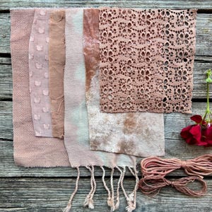 May include: A collection of fabric swatches in shades of dusty rose and beige, including a textured square with a grid pattern, a solid piece, a tie-dyed strip with fringe, a velvet-like piece, and a square of intricate floral lace, with two bundles of twine.