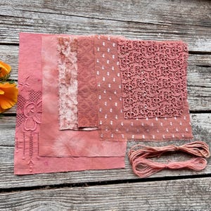 Puede incluir: Una colección de muestras de tela rosa con diversas texturas, incluyendo encaje, lunares y diseños bordados. También hay un ovillo de hilo rosa. Las telas están dispuestas sobre una superficie de madera envejecida, mostrando una estética rústica.