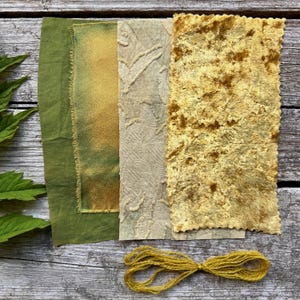 以下が含まれることがあります： 緑と金の色の生地見本を真上から見た写真。さまざまなテクスチャが特徴です。下部には黄色の糸の束があります。生地には、無地の緑、染めた緑と金、模様のあるベージュ、テクスチャード加工の金が含まれています。