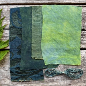 以下が含まれることがあります： 木製の表面に、さまざまな緑色の生地見本と緑色の糸の束が置かれています。生地は、濃いティールから明るいライムグリーンまで、質感と色合いが異なります。糸はくすんだティール色です。