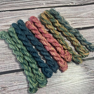 May include: Six skeins of colorful yarn in shades of green, teal, pink, and gold are arranged on a wooden surface. The yarn is tightly wound and appears soft, ideal for various crafting projects. The color variations add visual interest.