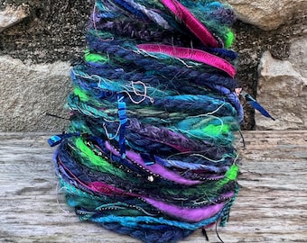 May include: A vibrant, hand-spun yarn bundle featuring a mix of textures and colors. The yarn includes shades of blue, green, purple, and pink, with ribbons and other embellishments woven throughout. This unique yarn is perfect for fiber art projects.