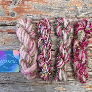 May include: Four colorful yarn skeins in shades of pink, green, and gold. The yarn is thick and textured, with a variety of fibers and embellishments. A card with the text "SUSTAINABLE FOSSICK FIBER LOVE" is in the lower left corner.
