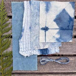 以下が含まれることがあります： インディゴ染めの様々なテキスタイルを真上から見た写真。無地の布、シワのある布、模様のある正方形の布が含まれています。青と白の糸の束が布の下にあります。木製の表面に配置されています。