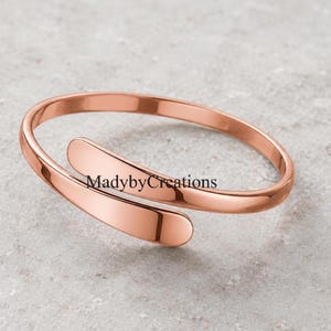 Könnte beinhalten: Ein roségoldfarbenes Bypass-Armband mit glatter, polierter Oberfläche. Das Metallband hat ein offenes Design mit zwei abgerundeten, sich überlappenden Enden. Der Text MadybyCreations ist deutlich auf der Oberfläche des Metallbandes zu sehen.
