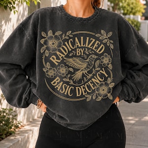 May include: A charcoal grey crewneck sweatshirt with a vintage wash finish. The front features a circular graphic in tan ink depicting a bird in flight surrounded by floral illustrations and the text RADICALIZED BY BASIC DECENCY.