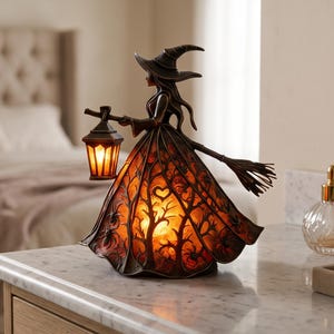 May include: A bronze-toned witch figurine lamp featuring a wide skirt illuminated from within, displaying a silhouette of bare trees and spiders. The witch wears a pointed hat, holds a glowing lantern in one hand, and carries a broomstick.