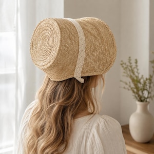 May include: A light tan straw bonnet featuring a cylindrical crown with a circular back panel and a wide brim. The hat is accented with a vertical strip of cream-colored lace trim that runs over the top and down the sides of the structure.