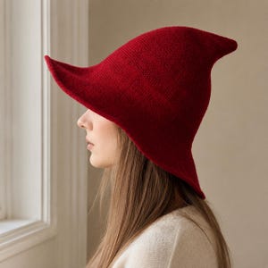 May include: A deep red knitted hat with a pointed crown and a wide, asymmetrical brim that curves upwards on one side. The hat features a fine, uniform knit texture and a structured shape that extends to a distinct point at the top.