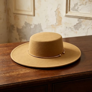 May include: A tan felt hat with a flat crown and a wide brim. The hat features a thin, metallic gold-toned circular band encircling the base of the crown, which is accented with two small, polished gold-toned spheres at the front opening.
