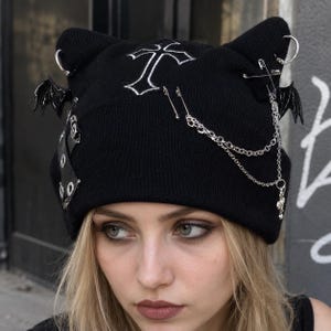 May include: A black knit beanie featuring cat-like ears, an embroidered white cross, two black bat-wing charms, silver-toned safety pins, metal rings, and a dangling silver chain. The design incorporates metal eyelets on a black strap attachment.