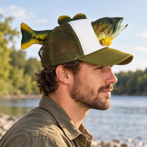 May include: A trucker hat featuring an olive green mesh back, a white front panel, and an olive green brim. A three-dimensional, realistic fish figure is attached to the top, with the head protruding from the front and the tail at the back.