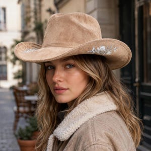 May include: A tan suede cowboy hat featuring a structured crown and a wide brim. The brim is decorated with intricate silver-toned floral embroidery on both sides. The hat has a smooth, matte texture and a classic curved shape along the edges.